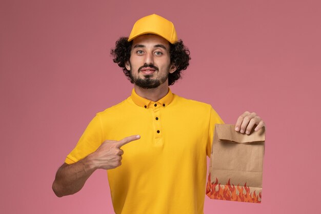 Männlicher Kurier der Vorderansicht in der gelben Uniform, die Papiernahrungsmittelpaket auf hellrosa Wand hält