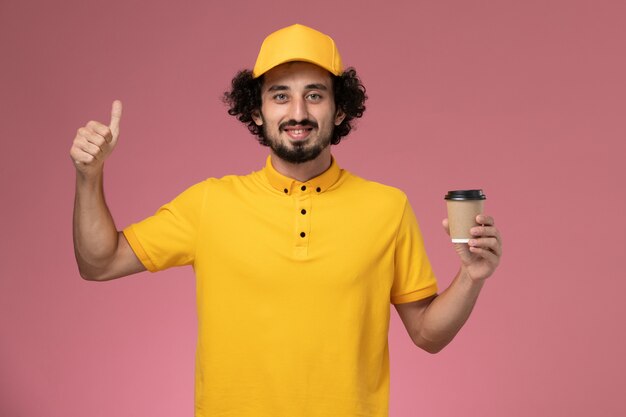 Männlicher Kurier der Vorderansicht in der gelben Uniform und im Umhang, der die Kaffeetasse der Lieferung an der rosa Wand hält