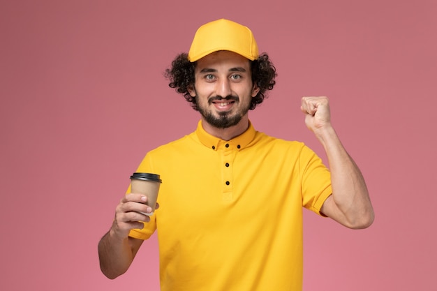 Männlicher Kurier der Vorderansicht in der gelben Uniform und im Umhang, der die Kaffeetasse der Lieferung an der rosa Wand hält