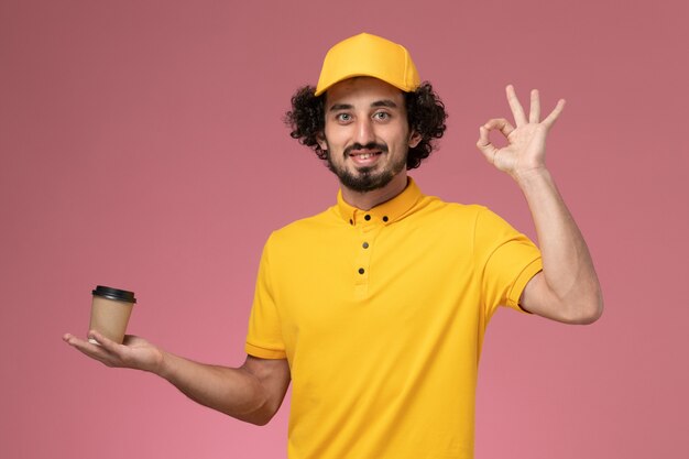 Männlicher Kurier der Vorderansicht in der gelben Uniform und im Umhang, der die Kaffeetasse der Lieferung an der rosa Wand hält