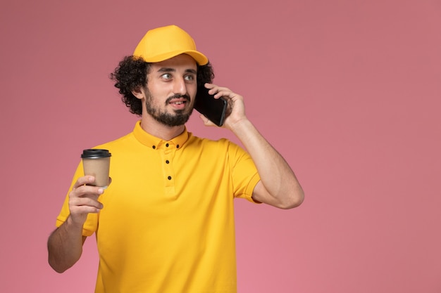 Männlicher Kurier der Vorderansicht in der gelben Uniform und im Umhang, der die Kaffeetasse und das Telefon der Lieferung an der rosa Wand hält