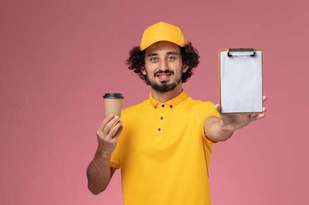 Männlicher Kurier der Vorderansicht in der gelben Uniform und im Umhang, der die Kaffeetasse und den Notizblock der Lieferung an der rosa Wand hält