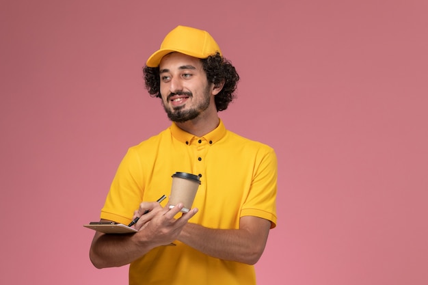 Männlicher Kurier der Vorderansicht in der gelben Uniform und im Umhang, der Lieferung Kaffeetasse Stift und Notizblock auf rosa Wand hält