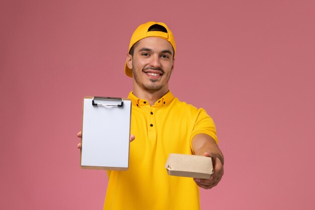 Männlicher Kurier der Vorderansicht in der gelben Uniform und im Umhang, der Notizblock des kleinen Lieferlebensmittelpakets auf rosa Hintergrund hält.