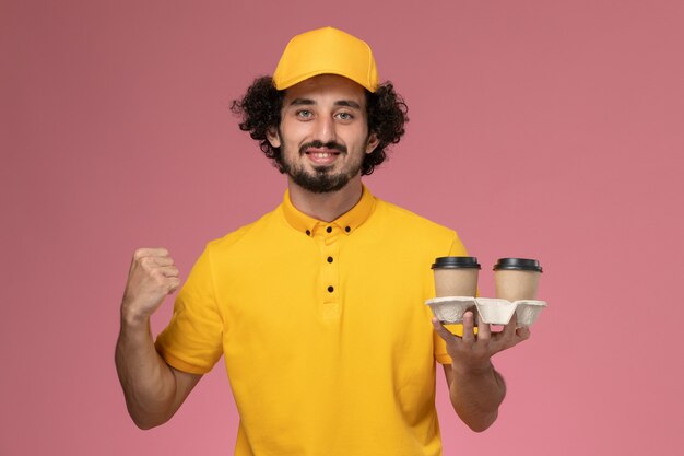 Männlicher Kurier der Vorderansicht in der gelben Uniform und im Umhang, die braune Kaffeetassen der Lieferung halten, die auf rosa Wand jubeln
