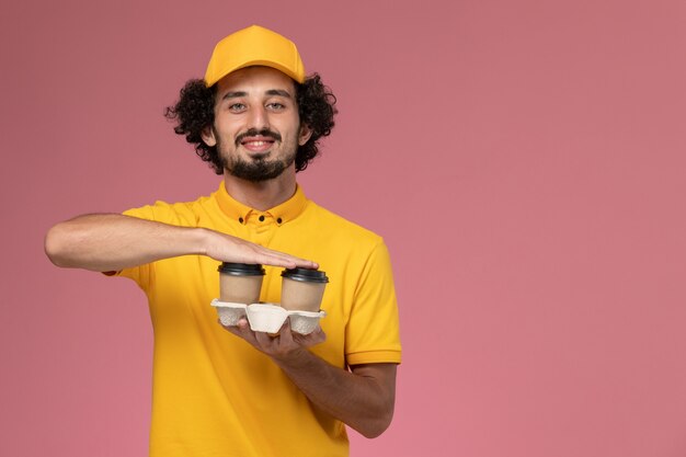 Männlicher Kurier der Vorderansicht in der gelben Uniform und im Umhang, die braune Lieferkaffeetassen mit einem Lächeln auf der rosa Wand halten