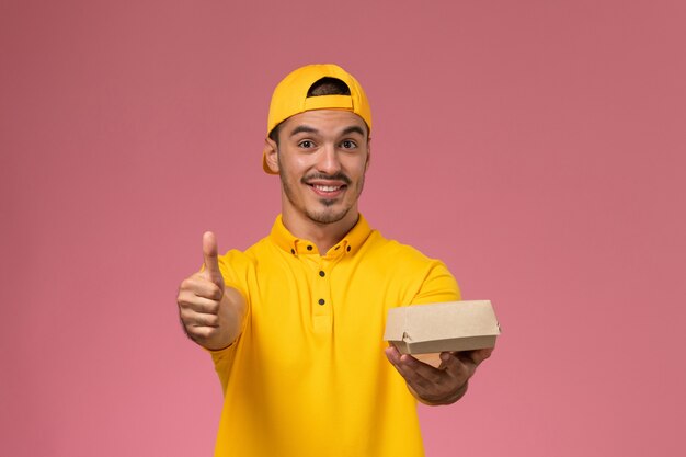 Männlicher Kurier der Vorderansicht in der gelben Uniform und im Umhang, die kleines Liefernahrungsmittelpaket mit Lächeln auf dem rosa Hintergrund halten.