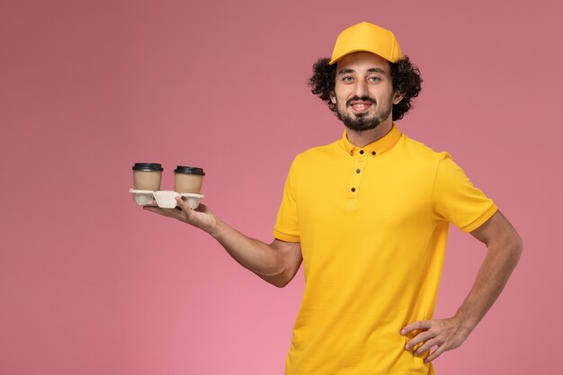 Männlicher Kurier der Vorderansicht in der gelben Uniform und im Umhang, die Lieferkaffeetassen auf rosa Wand halten