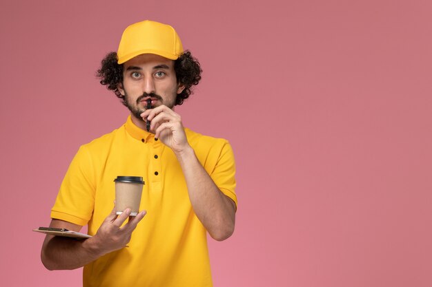 Männlicher Kurier der Vorderansicht in der gelben Uniform und im Umhang, die Lieferung Kaffeetasse Stift und Notizblock denken auf rosa Wand halten