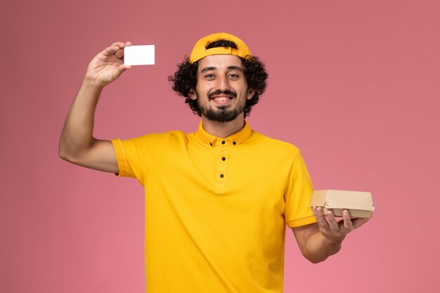 Männlicher Kurier der Vorderansicht in der gelben Uniform und im Umhang mit Karte und kleinem Liefernahrungsmittelpaket auf seinen Händen auf dem hellrosa Hintergrund.