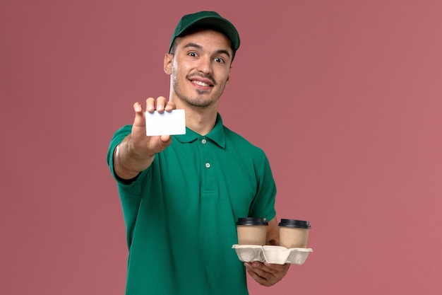 Männlicher Kurier der Vorderansicht in der grünen Uniform, die braune Kaffeetassen der Lieferung und Plastikkarte auf rosa Schreibtischmann hält