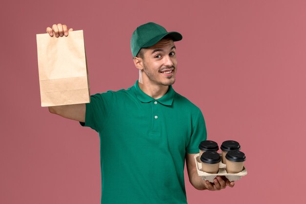 Männlicher Kurier der Vorderansicht in der grünen Uniform, die braune Kaffeetassen und Nahrungspaket verpackt, die auf rosa Hintergrund lächeln