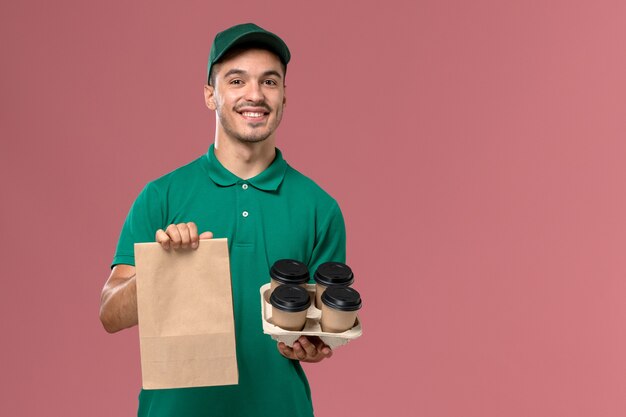 Männlicher Kurier der Vorderansicht in der grünen Uniform, die braune Kaffeetassen und Nahrungspaket verpackt, die auf rosa Hintergrund lächeln