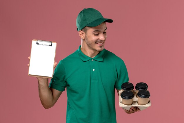 Männlicher Kurier der Vorderansicht in der grünen Uniform, die braune Kaffeetassen und Notizblock lächelnd auf rosa Hintergrund hält