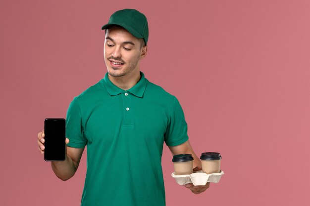 Männlicher Kurier der Vorderansicht in der grünen Uniform, die braune Kaffeetassen und Telefon der Lieferung auf dem rosa Schreibtisch hält