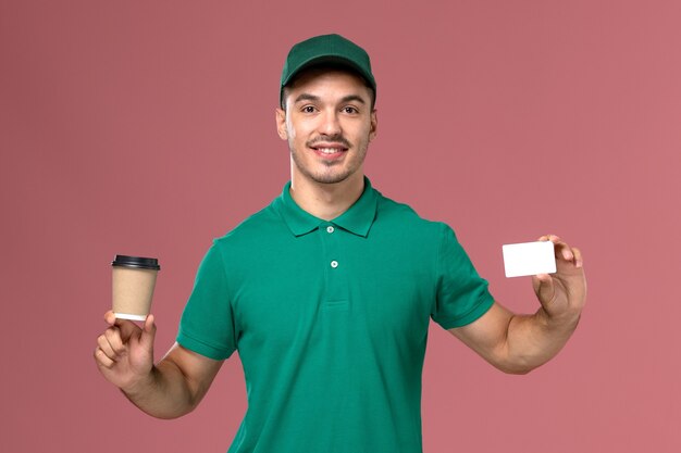 Männlicher Kurier der Vorderansicht in der grünen Uniform, die die Kaffeetasse der Lieferung mit weißer Karte auf dem hellrosa Hintergrund hält