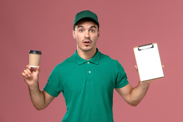 Männlicher Kurier der Vorderansicht in der grünen Uniform, die die Kaffeetasse und den Notizblock der Lieferung auf dem rosa Schreibtisch hält