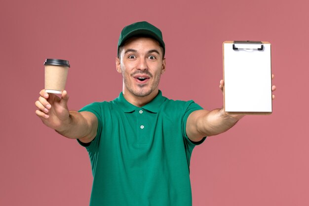 Männlicher Kurier der Vorderansicht in der grünen Uniform, die die Kaffeetasse und den Notizblock hält, die sich auf hellrosa Schreibtisch freuen