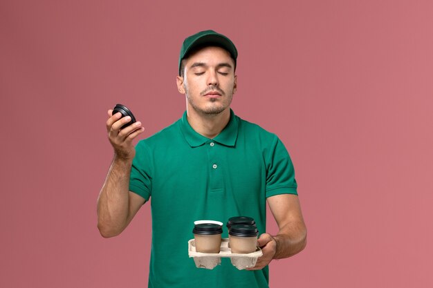 Männlicher Kurier der Vorderansicht in der grünen Uniform, die Kaffeetassen der Lieferung hält, die ihren Duft auf rosa Schreibtisch riechen