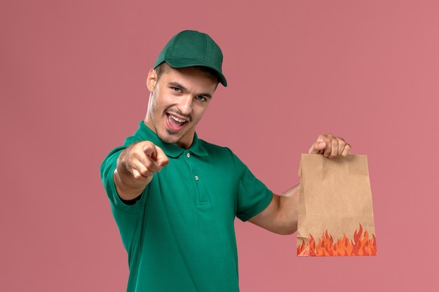 Männlicher Kurier der Vorderansicht in der grünen Uniform, die Papiernahrungsmittelpaket hält und auf hellrosa Hintergrund hinweist