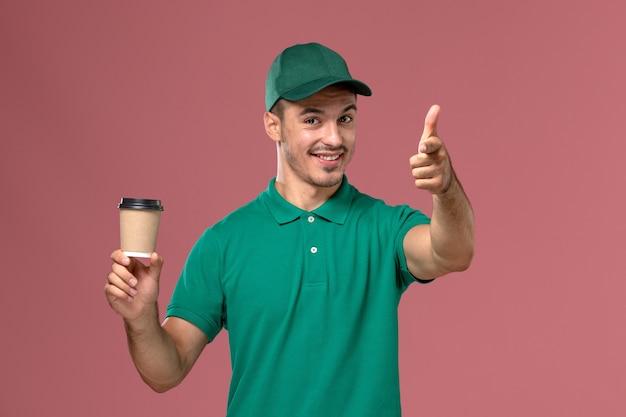 Männlicher Kurier der Vorderansicht in der grünen Uniform lächelnd und Kaffeetasse auf dem rosa Hintergrund haltend