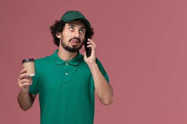 Männlicher Kurier der Vorderansicht in der grünen Uniform und im Umhang, die Kaffeetasse halten und am Telefon auf dem rosa Hintergrunduniformlieferdienst sprechen