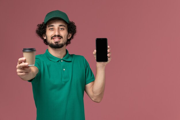 Männlicher Kurier der Vorderansicht in der grünen Uniform und im Umhang, die Kaffeetasse mit Telefon auf rosa Hintergrunduniform-Lieferauftragsservice halten