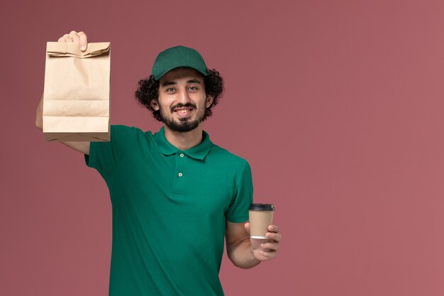 Männlicher Kurier der Vorderansicht in der grünen Uniform und im Umhang, die Kaffeetasse und Lebensmittelpaket der Lieferung auf rosa einheitlichem Lieferserviceunternehmen des Hintergrunds halten