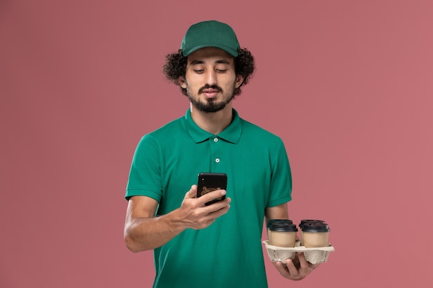 Männlicher Kurier der Vorderansicht in der grünen Uniform und im Umhang, die Kaffeetassen mit Telefon auf hellrosa Hintergrunddienstuniformlieferarbeiterjob halten