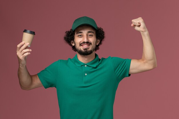 Männlicher Kurier der Vorderansicht in der grünen Uniform und im Umhang, die Lieferkaffeetasse halten und auf dem rosa Hintergrunduniformlieferdienstfirmenjobarbeiter biegen