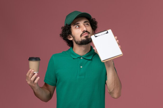 Männlicher Kurier der Vorderansicht in der grünen Uniform und im Umhang, die Lieferung Kaffeetasse mit Notizblock denken auf dem rosa Hintergrunduniformlieferdienstfirmenjob denken