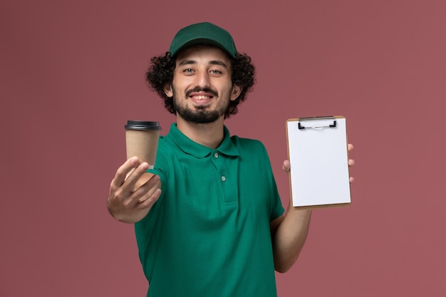 Männlicher Kurier der Vorderansicht in der grünen Uniform und im Umhang, die Lieferung Kaffeetasse mit Notizblock und Lächeln auf rosa Schreibtischuniformlieferdienstfirmenjob halten