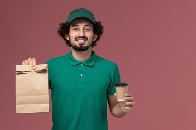 Männlicher Kurier der Vorderansicht in der grünen Uniform und im Umhang, die Lieferung Kaffeetasse und Lebensmittelpaket auf rosa Schreibtischuniform-Lieferservice-Job halten