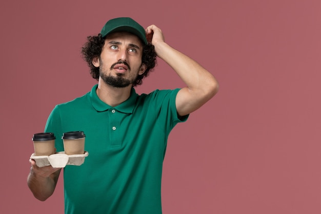 Männlicher Kurier der Vorderansicht in der grünen Uniform und im Umhang, die Lieferung Kaffeetassen halten, die auf rosa Hintergrunddienstuniformlieferungsarbeitsarbeit denken
