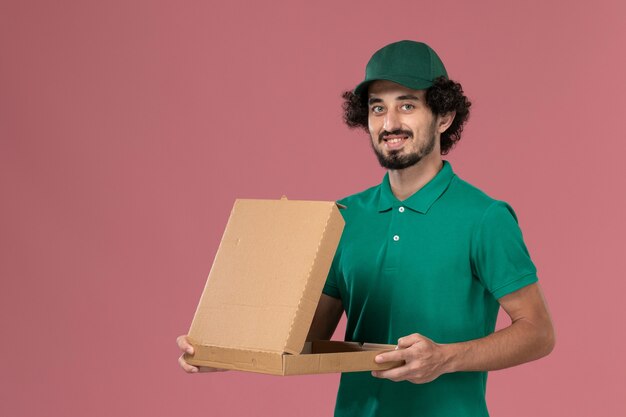 Männlicher Kurier der Vorderansicht in der grünen Uniform und im Umhang, die Nahrungsmittelbox auf rosa Hintergrunddienstarbeiteruniformlieferarbeitsmann halten