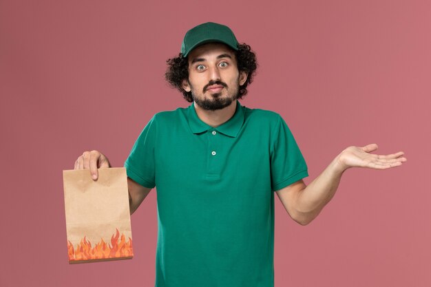 Männlicher Kurier der Vorderansicht in der grünen Uniform und im Umhang, die Papiernahrungsmittelpaket halten und mit verwirrtem Ausdruck auf rosa Hintergrunddienstuniformlieferauftrag aufwerfen