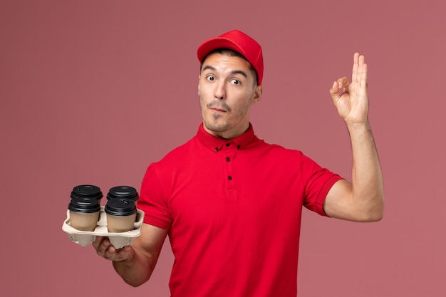 Männlicher Kurier der Vorderansicht in der roten Uniform, die braune Kaffeetassen der Lieferung auf dem hellrosa Wandarbeiter hält