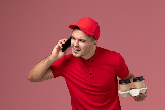 Männlicher Kurier der Vorderansicht in der roten Uniform, die braune Kaffeetassen der Lieferung hält und am Telefon auf rosa Schreibtisch spricht