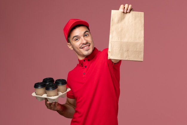 Männlicher Kurier der Vorderansicht in der roten Uniform, die braune Kaffeetassen der Lieferung mit Lebensmittelverpackung an der rosa Wand hält