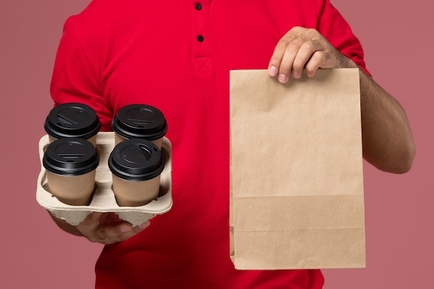 Kostenloses Foto männlicher kurier der vorderansicht in der roten uniform, die braune kaffeetassen der lieferung mit lebensmittelverpackung auf der rosa wanddienst-zustelljobarbeiteruniform hält