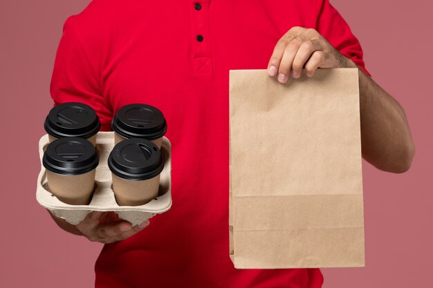 Männlicher Kurier der Vorderansicht in der roten Uniform, die braune Kaffeetassen der Lieferung mit Lebensmittelverpackung auf der rosa Wanddienst-Zustelljobarbeiteruniform hält