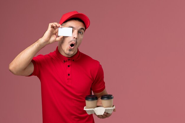 Männlicher Kurier der Vorderansicht in der roten Uniform, die braune Kaffeetassen der Lieferung mit weißer Karte auf der rosa Wand hält