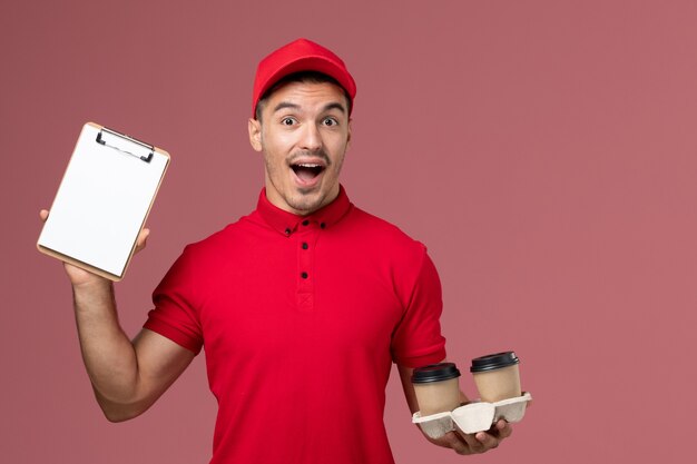 Männlicher Kurier der Vorderansicht in der roten Uniform, die braune Kaffeetassen und Notizblock der Lieferung auf hellrosa Schreibtischarbeiter hält