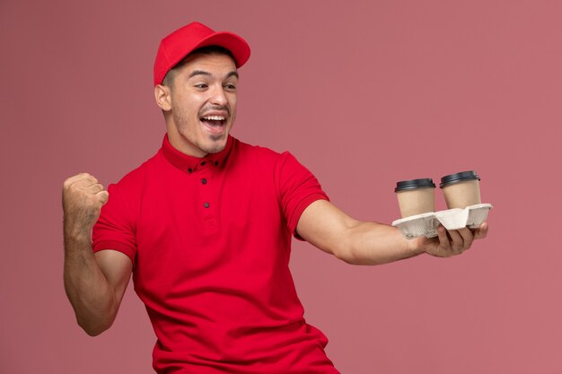 Männlicher Kurier der Vorderansicht in der roten Uniform, die braune Lieferkaffeetassen hält, die sich auf rosa Wand freuen