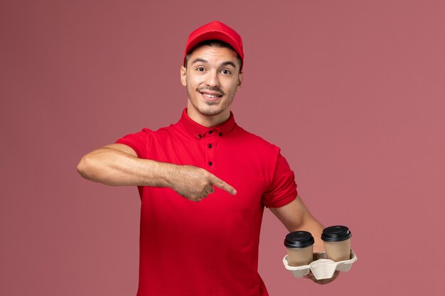 Männlicher Kurier der Vorderansicht in der roten Uniform, die braune Lieferung Kaffeetasse auf rosa Wand Service Lieferung männliche Uniform Job hält