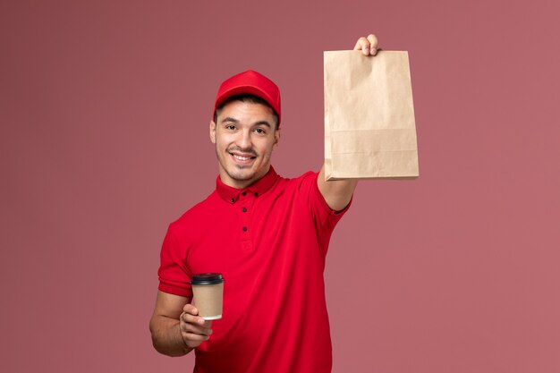 Männlicher kurier der vorderansicht in der roten uniform, die die kaffeetasse der lieferung und das lebensmittelpaket mit dem lächeln auf dem männlichen uniformjob des rosa wanddienstlieferarbeiters hält