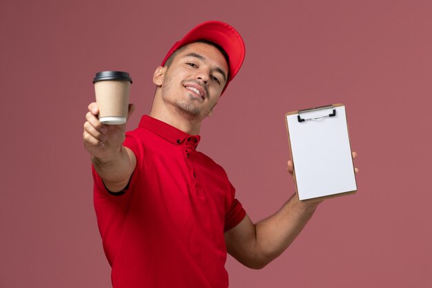 Männlicher Kurier der Vorderansicht in der roten Uniform, die die Kaffeetasse und den Notizblock der Lieferung auf dem rosa Wandarbeiter männlich hält