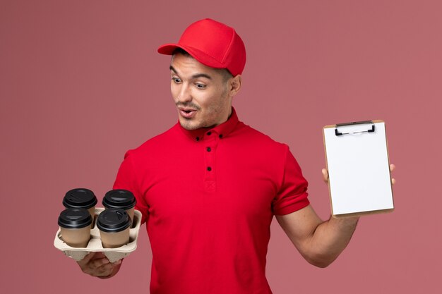 Männlicher Kurier der Vorderansicht in der roten Uniform, die Kaffeetassen der Lieferung mit Notizblock auf rosa Wand hält