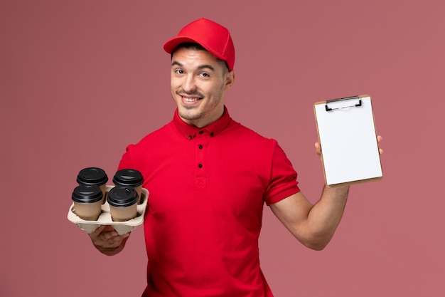 Männlicher Kurier der Vorderansicht in der roten Uniform, die Kaffeetassen der Lieferung mit Notizblock auf rosa Wand hält