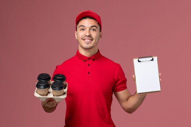Männlicher Kurier der Vorderansicht in der roten Uniform, die Kaffeetassen der Lieferung mit Notizblocklächeln auf rosa Wand hält